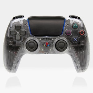 Puede incluir: Un mando inalámbrico PlayStation 5 DualSense transparente con botones negros y un acento azul. El mando tiene un logotipo de PlayStation en la parte delantera.