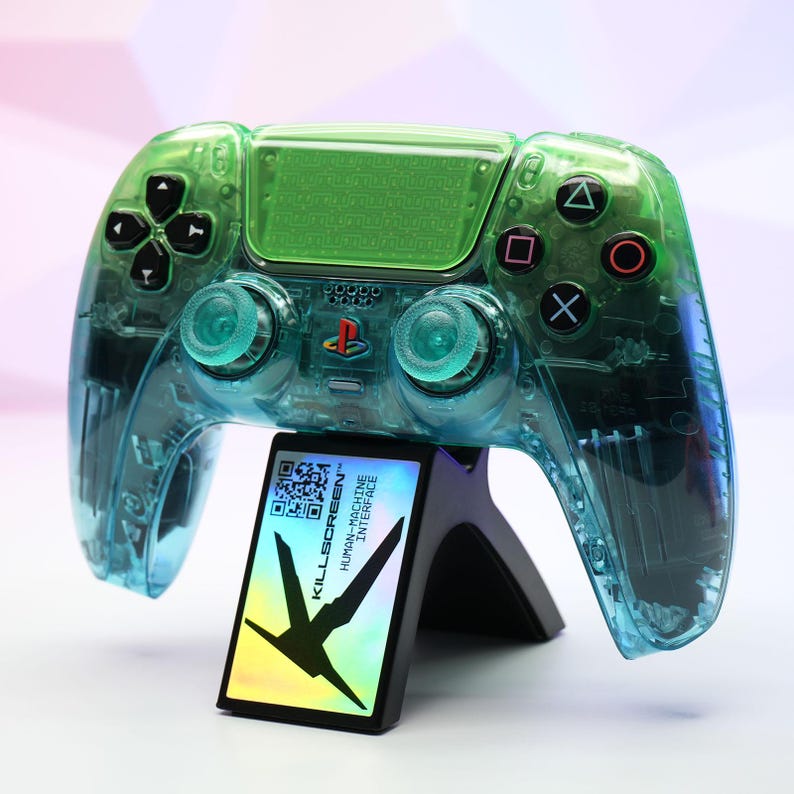 Peut inclure: Une manette de jeu vid&eacute;o transparente verte et bleue avec des boutons noirs et un support. La manette a un design clair, mettant en valeur les composants internes. Le support affiche le texte "KILLSCREEN HUMAN-MACHINE INTERFACE".
