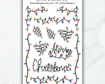 May include: A sheet of Christmas themed stickers featuring the words "Merry Christmas" and "Cheer" written in a script font with colorful string lights. The stickers are on a white background.