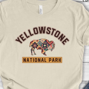 May include: A beige t-shirt with a brown and orange graphic of a bison with a colorful geometric pattern. The text "Yellowstone National Park" is printed in brown letters below the bison.