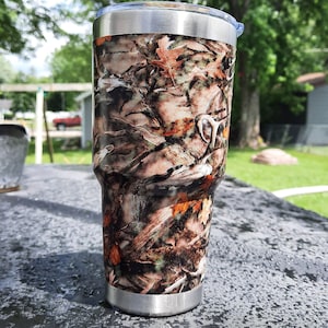 May include: A stainless steel tumbler with a camouflage pattern featuring brown, orange, and green hues. The tumbler has a silver rim and a clear lid. A white logo is visible on the side. The tumbler is designed to keep drinks hot or cold.
