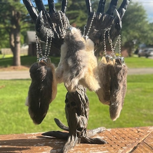 May include: A dark, gothic-style sculpture featuring a hand-like structure with attached animal paws. The paws are various shades of brown and gray, suspended by silver chains. The sculpture is set outdoors on a wooden surface.