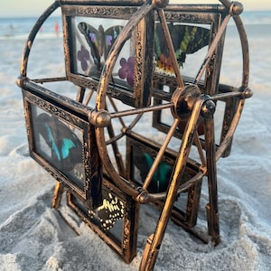 May include: A bronze-colored metal ferris wheel display stand featuring multiple rectangular frames. Each frame contains a preserved butterfly specimen with vibrant wings and pressed purple flowers, encased behind clear glass panels.