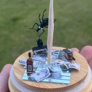 May include: A miniature scene with a blue beetle on a white stick, surrounded by dollar bills, a bottle of whiskey, and a mirrored surface on a wooden base.
