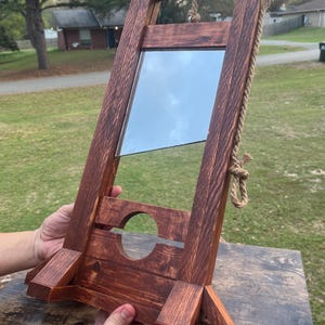 May include: A wooden guillotine replica featuring a dark brown stained finish, a reflective metal blade suspended by a thick jute rope, and a base with a circular cutout. The structure is supported by angled wooden feet on both sides.