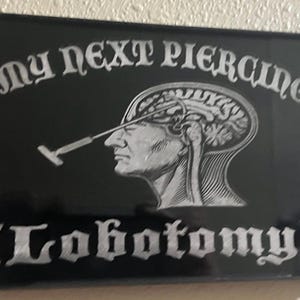 May include: Black rectangular sign with the words "MY NEXT PIERCING" in an ornate font. Below is a detailed illustration of a human head with a brain and a surgical tool. The word "(Lobotomy)" is below the illustration.