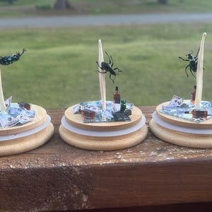 May include: Three miniature wooden bases with a white stick and a black beetle on top. Each base has a pile of fake money and a small bottle on top. The bases are on a wooden surface.