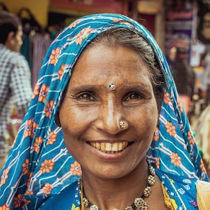 May include: A woman wearing a blue and orange patterned headscarf smiles at the camera. She is wearing a colorful patterned blouse and a silver necklace with dangling charms.