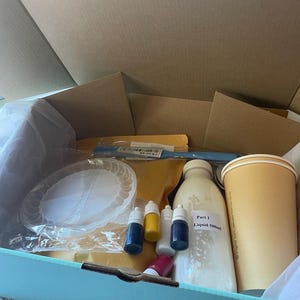 May include: An open box containing craft supplies. Includes a bottle labelled "Part 1 Liquid 500ml", small bottles of coloured liquids, a paper plate, a stirring stick, and a paper cup. The box is light blue.