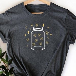 May include: Dark gray t-shirt featuring a white illustration of a jar filled with fireflies. The design includes gold-colored stars and diamond shapes around the jar. The shirt is displayed on a wooden hanger.