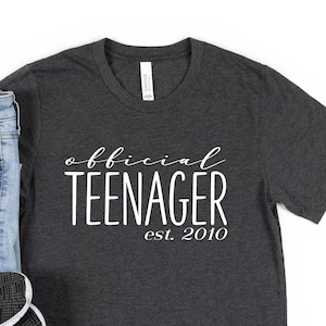 May include: A dark grey t-shirt with the text "official teenager est. 2010" in white. The shirt is paired with blue jeans and black trainers.