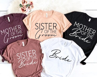 May include: A collection of five short-sleeved t-shirts in various colors, including burgundy, peach, and black. Each shirt features a different title in white script: "Mother of the Groom," "Sister of the Groom," "Mother of the Bride," "Sister of the Bride," and "Bride."