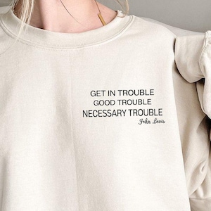 May include: A beige sweatshirt with the text "GET IN TROUBLE GOOD TROUBLE NECESSARY TROUBLE John Lewis" printed in black. The sweatshirt is a neutral color and has long sleeves. The text is centered on the chest.