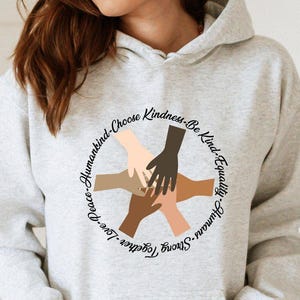May include: A light gray hoodie with a front pocket and a drawstring hood. The graphic on the front features a circle of hands in various skin tones, with the words "Peace, Love, Together, Strong, Humans, Choose Kindness, Be Kind, Equally" around the circle.