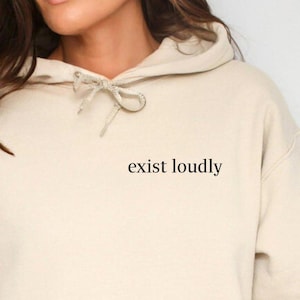 May include: A beige hoodie with a front pocket and drawstring hood. The phrase "exist loudly" is printed in a small, black font on the upper chest area of the hoodie. The hoodie is made of a soft, comfortable material.