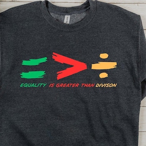 May include: Dark gray sweatshirt with the text "EQUALITY IS GREATER THAN DIVISION". The graphic features green equal signs, a red greater-than symbol, and an orange division symbol. The sweatshirt is a crew neck.