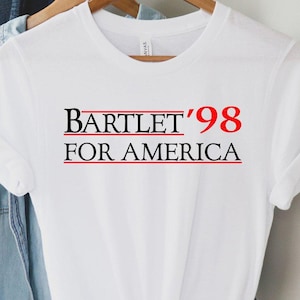 May include: White t-shirt with the text "BARTLET '98 FOR AMERICA" in black and red. The shirt is hanging on a wooden hanger with a denim shirt and a brown hat in the background.