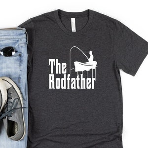 May include: A dark heather grey t-shirt with a white graphic of a person fishing from a boat. The text "The Rodfather" is printed below the graphic.