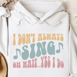 May include: White hoodie with the text "I don't always sing, oh wait... yes I do" in retro-style lettering. The text is in shades of orange, blue, and pink. The hoodie has a drawstring hood and long sleeves.