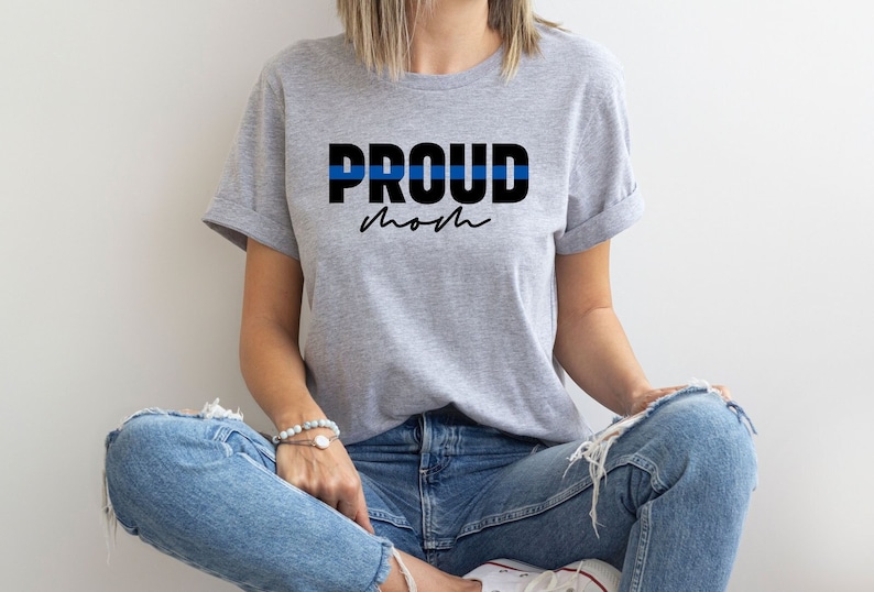 以下が含まれることがあります： 「PROUD mom」という黒い文字が書かれたグレーのTシャツ。PROUDの上には細い青い線が引かれている。