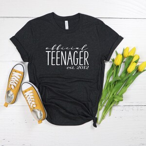 May include: A dark grey t-shirt with the text "official teenager est. 2012" in white lettering. The shirt is lying on a white surface with yellow flowers.
