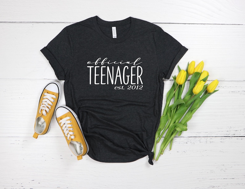 May include: A dark grey t-shirt with the text "official teenager est. 2012" in white lettering. The shirt is lying on a white surface with yellow flowers.