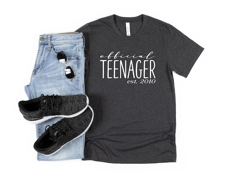 May include: A dark grey t-shirt with the text "official teenager est. 2010" in white. The shirt is paired with blue jeans and black trainers.