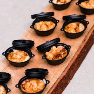 May include: A wooden serving board with small black cast iron pots filled with shrimp and grits. The pots have lids.