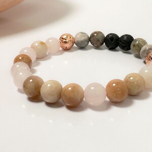 Op de afbeelding: Een armband met roze, bruine en grijze kralen, en twee koperkleurige kralen. De armband ligt op een wit oppervlak.