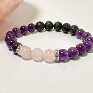 May include: A purple and pink gemstone bracelet with black lava beads. The bracelet has a silver charm with rhinestones.