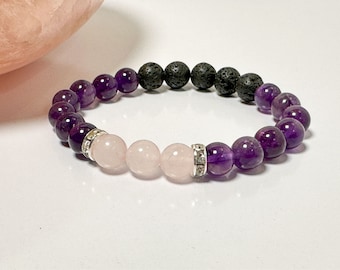 May include: A purple and pink gemstone bracelet with black lava beads. The bracelet has a silver charm with rhinestones.