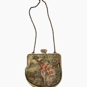 Puede incluir: Bolso de mano vintage de tapiz con marco y cadena de metal dorado. El bolso presenta una escena pastoral en tonos verdes, beige y rojos. El cierre es ornamentado y el bolso tiene una base curva.