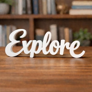 May include: A white wooden sign that spells out the word "Explore" in a cursive font. The sign is placed on a wooden surface, with a blurred background of a bookshelf filled with books.