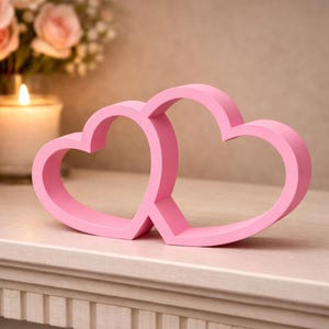 May include: A light pink, hollow, double-heart shaped decorative object. The piece features two interconnected heart outlines with smooth, flat surfaces and rounded edges, standing upright as a singular, unified structure on a flat surface.