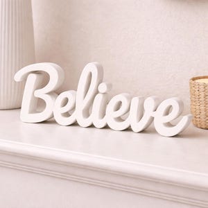 May include: A white, freestanding decorative sign featuring the word Believe in a cursive, script-style font. The letters are connected in a single piece, with a smooth, matte finish, designed to stand upright on a flat surface for display.