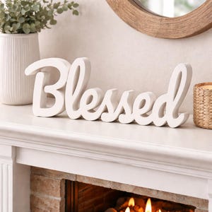 Pode incluir: Uma placa de madeira branca com a palavra "Blessed" escrita em itálico, colocada numa lareira branca. Um vaso com vegetação e uma vela num suporte tecido são visíveis no fundo. A lareira está acima de uma lareira de tijolo.