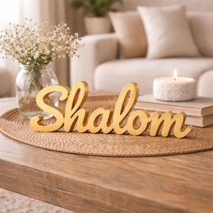 May include: A gold-coloured 'Shalom' sign sits on a woven mat on a wooden table. A vase of white flowers and a lit candle are nearby. The scene is set in a cosy living room.