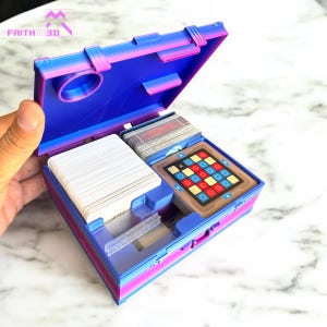 以下が含まれることがあります： 青とピンクの3Dプリントされたゲームケースで、ゲームコンポーネントが表示されています。ケースにはカードの山と、色の付いたタイルが付いたゲーム盤が入っています。ケースにはハンドルと留め金が付いています。「FAITH 3D」の文字が見えます。