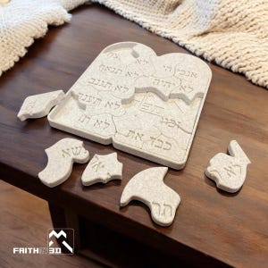 May include: A white wooden puzzle shaped like a tablet with Hebrew text. The puzzle is made up of several pieces, some of which are scattered around the main piece. The text on the puzzle reads: "אנכי ה' אלהיך לא יהיה לא תנאף לא תגנב לא תענה כבד את אביך ואמך לא תרצח לא תשא את שם ה' אלהיך לשוא זכור את יום השבת לקדשו ששת ימים תעבוד ועשית כל מלאכתך יום השביעי שבת לה' אלהיך לא תעשה כל מלאכה אתה ובנך ובתך עבדך ואמתך בהמתך וגרך אשר בשעריך כי בששת ימים עשה ה' את השמים ואת הארץ את הים ואת כל אשר בם וינפש ביום השביעי לכן ברך ה' את יום השבת וקדשו".