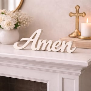 May include: A white wooden cutout sign featuring the word Amen in a cursive script font. The letters are connected in a single piece, designed to stand upright on a flat surface. The finish is smooth and matte, providing a clean appearance.