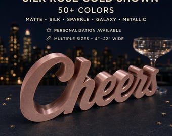 May include: A rose gold, three-dimensional script sign reading 'Cheers'. Text above states: 'SILK ROSE GOLD SHOWN, 50+ COLORS, MATTE, SILK, SPARKLE, GALAXY, METALLIC, PERSONALIZATION AVAILABLE, MULTIPLE SIZES, 4"–22" WIDE'. The sign has a smooth finish.