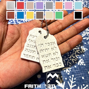 Könnte beinhalten: Zwei weiße, steinartige Anhänger mit hebräischem Text, die an einer silbernen Kette hängen. Die Anhänger sind rechteckig geformt wie Hundemarken. Der Text auf den Anhängern lautet: "אנכי ה' אלקיך לא יהיה לך אלהים אחרים" und "לא תעשה לך פסל וכל תמונה אשר בשמים"