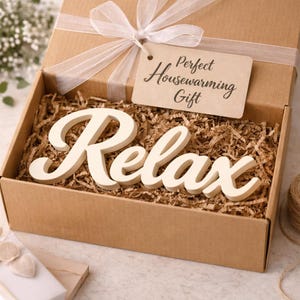 May include: A gift box with the word "Relax" in cream-colored script. The box is filled with brown paper and tied with a white ribbon. A tag reads "Perfect Housewarming Gift".