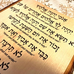 Op de afbeelding: Een houten plaquette met de Tien Geboden in het Hebreeuws. De tekst is in het hout gebrand en is gemakkelijk te lezen. De plaquette is lichtbruin van kleur en heeft een natuurlijke houtnerf.