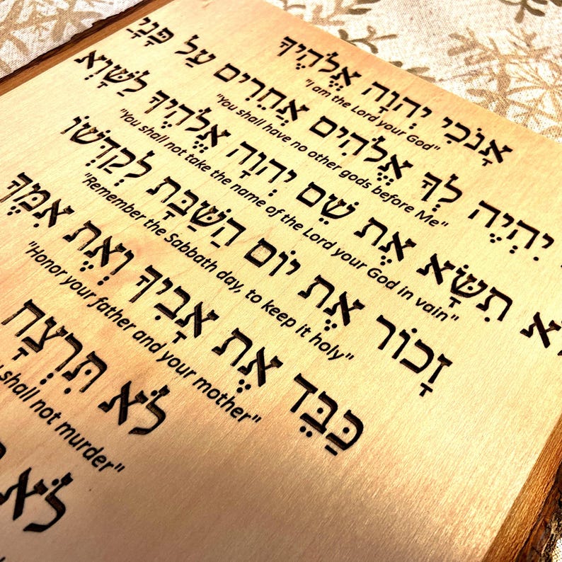 Op de afbeelding: Een houten plaquette met de Tien Geboden in het Hebreeuws. De tekst is in het hout gebrand en is gemakkelijk te lezen. De plaquette is lichtbruin van kleur en heeft een natuurlijke houtnerf.