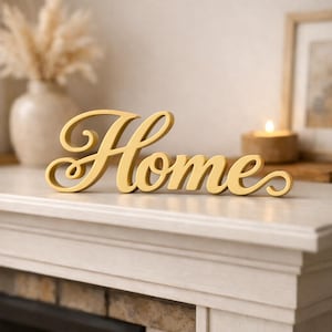 May include: A gold-coloured, freestanding decorative sign featuring the word Home in a cursive, flowing script font. The letters are connected in a single piece, with a smooth finish and rounded edges, designed to sit on a flat surface.