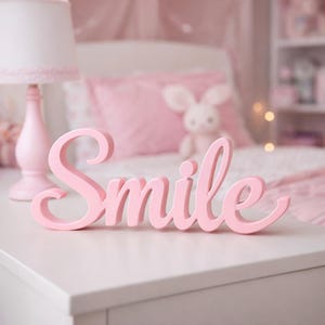 May include: A light pink, freestanding wooden sign featuring the word Smile in a cursive, flowing script font. The letters are connected in a single piece, with a smooth, matte finish, designed to sit upright on a flat surface.