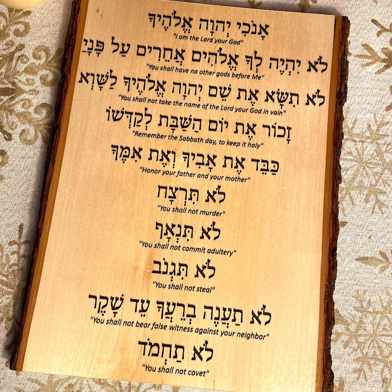 Op de afbeelding: Een houten plaquette met de Tien Geboden in het Hebreeuws en Engels. De plaquette is lichtbruin met een natuurlijke houtnerf. De tekst is gegraveerd in donkerbruin.