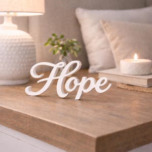 Puede incluir: Un letrero de madera blanco independiente que muestra la palabra Hope en una fuente cursiva. Las letras están conectadas y tienen un acabado mate suave, diseñado para mantenerse en posición vertical sobre una superficie plana.