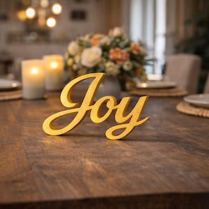 Może przedstawiać: Złoty napis "Joy" w kursywie umieszczony na ciemnym drewnianym stole. Słowo jest centralnym punktem, z rozmytym tłem świec, kwiatów i ustawienia jadalni. Słowo jest dekoracyjnym akcentem do wystroju domu.